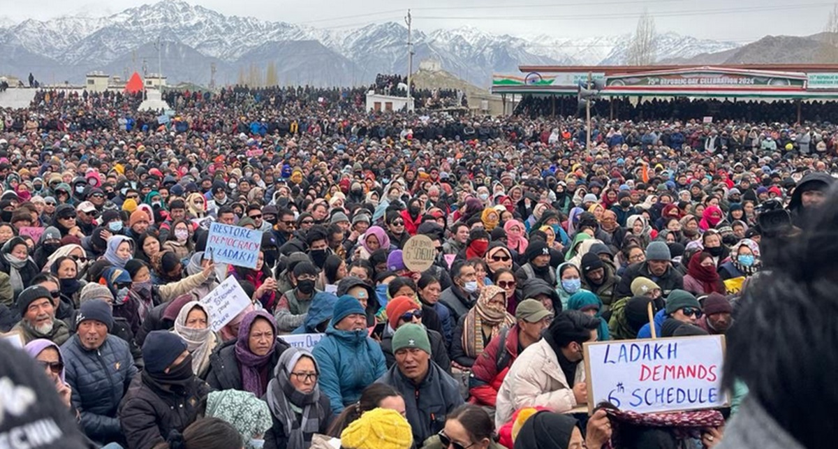 A massive crowd in Leh, Ladakh during a protest demanding statehood and inclusion under the Sixth Schedule, with people holding placards like “Restore Democracy in Ladakh” and “Ladakh Demands 6th Schedule” against the backdrop of snow-covered mountains.